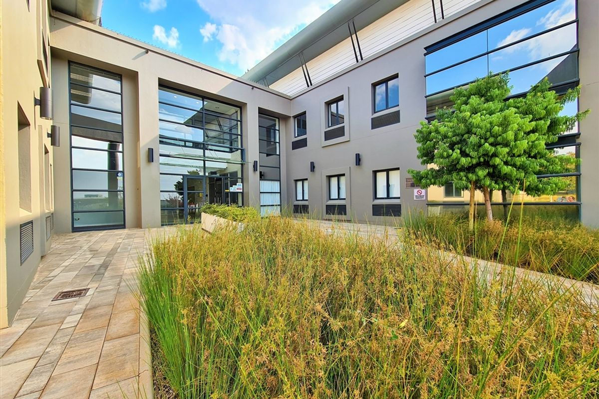 Modern office building exterior featuring a landscaped courtyard, paved walkways, and extensive glass facade elements.