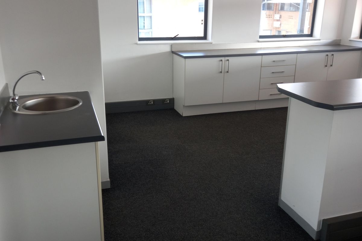 A functional office kitchenette area featuring built-in cabinetry, a stainless steel sink, and dark countertops, set against a neutral office backdrop.