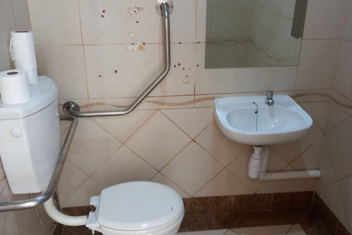 A functional office restroom featuring a wall-mounted basin, toilet, and stainless steel grab rails with tiled walls and flooring.
