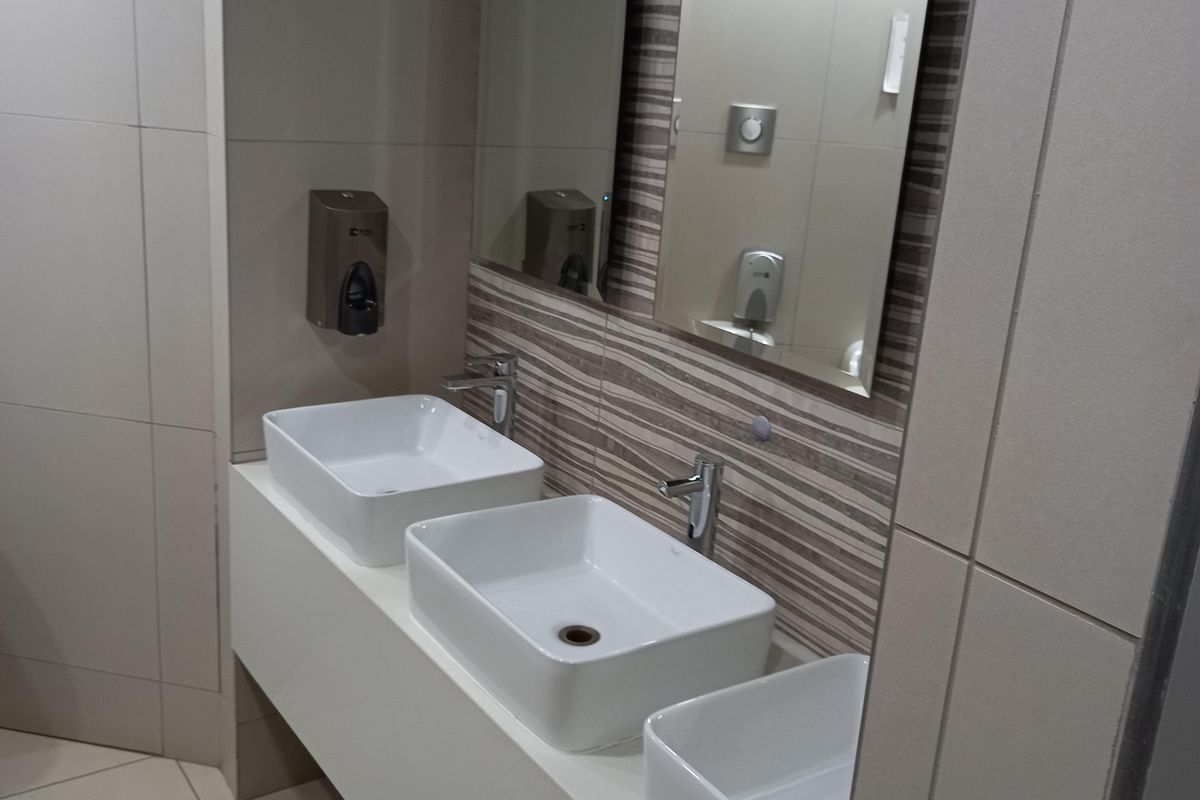 Modern office restroom featuring a sleek white vanity with three vessel sinks, chrome faucets, and decorative tiled wall accents with integrated mirrors.