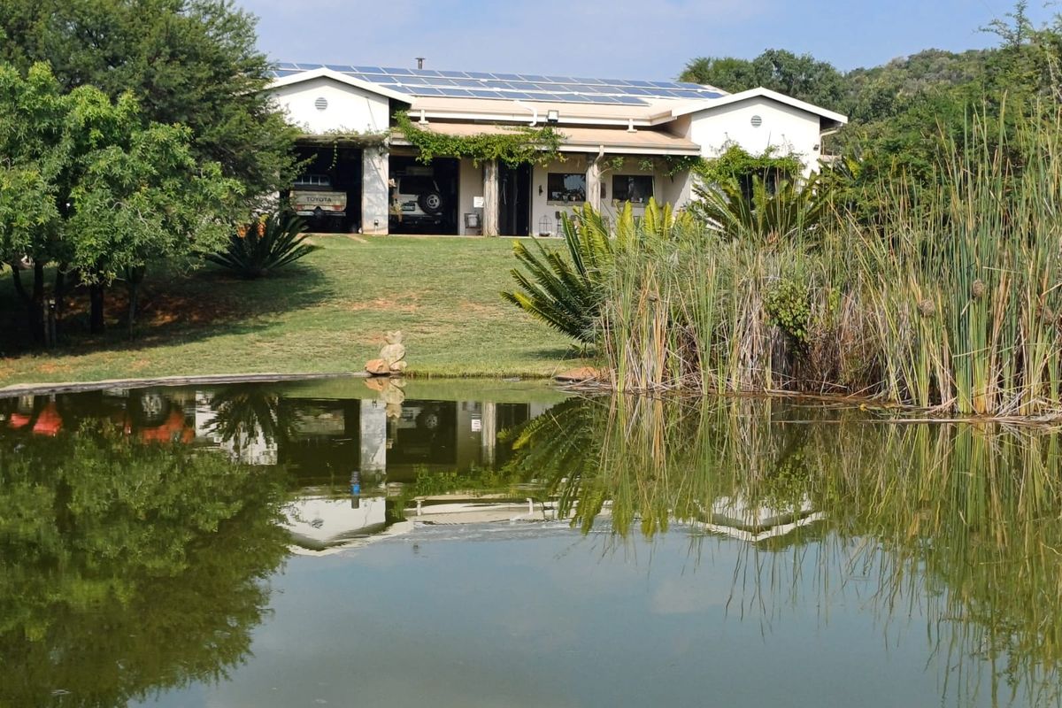 A scenic view of the rear farm house elevation featuring a covered patio, solar panel installation, and a tranquil pond in the foreground.