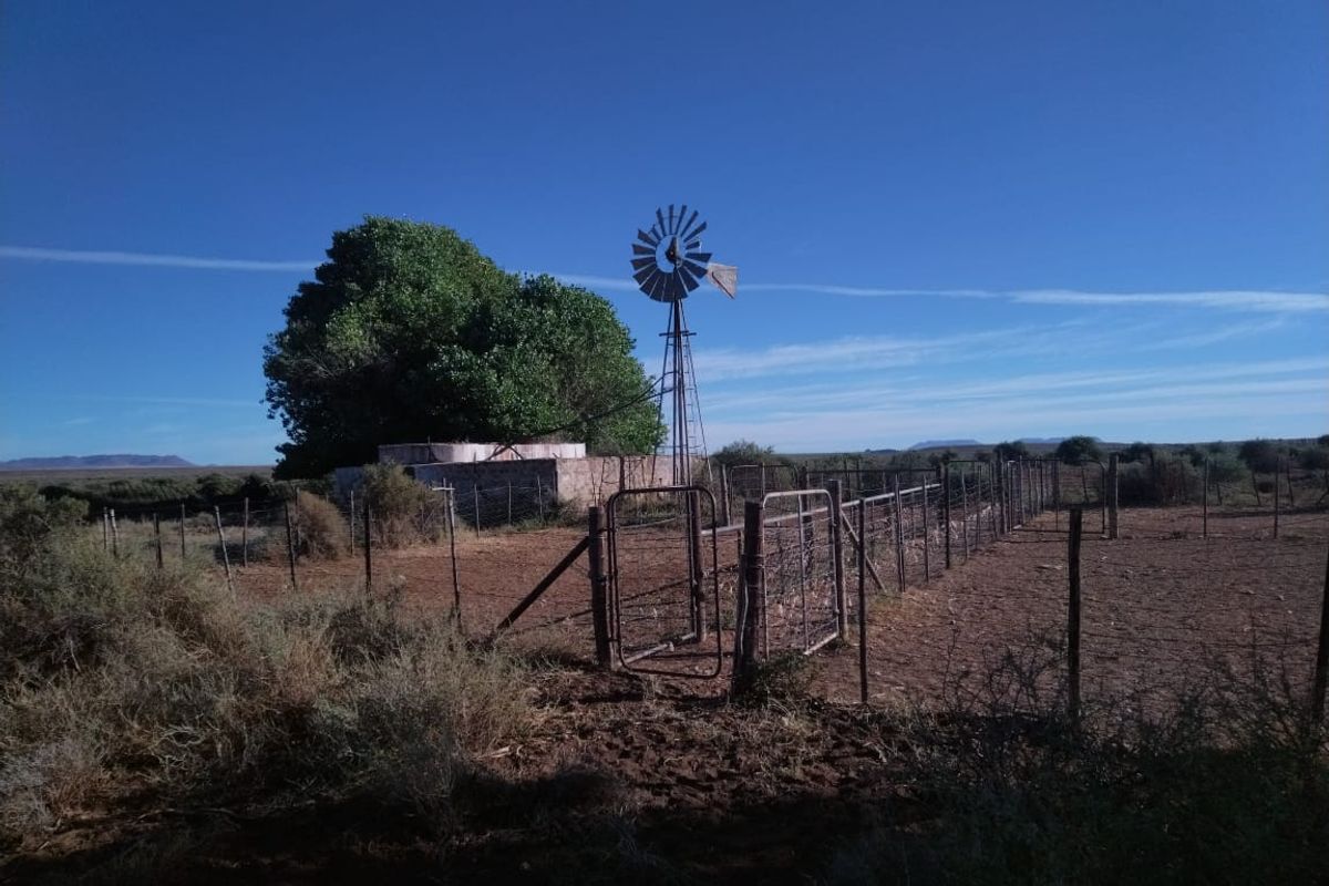 A rural farm scene featuring a traditional windmill, a water reservoir, and extensive wire fencing across the open landscape.
