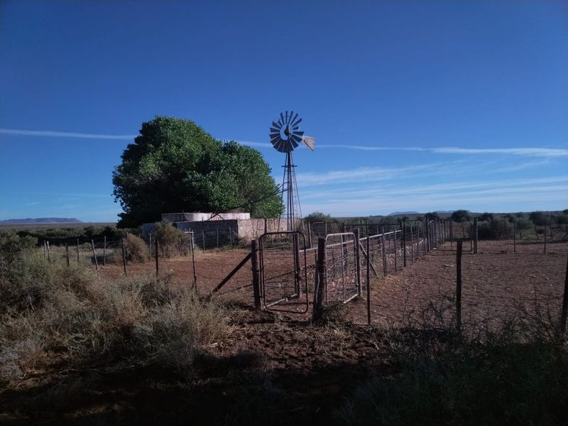 A rural farm scene featuring a traditional windmill, a water reservoir, and extensive wire fencing across the open landscape.