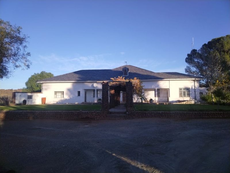 Front elevation of a farm-style residence featuring a stone-clad pergola entrance, white exterior walls, and a tiled roof set against a gravel driveway.