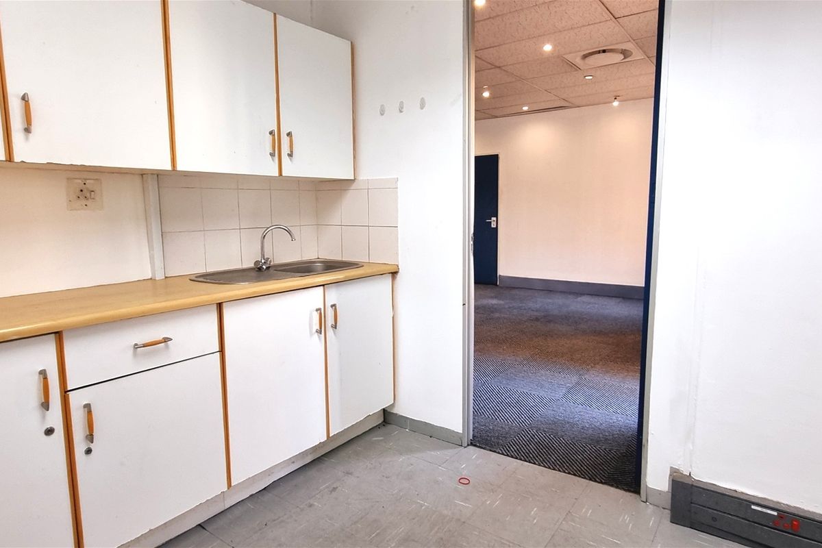 A functional office kitchenette featuring white cabinetry, a laminate countertop, and a stainless steel sink, leading into an open-plan office space with carpeted flooring.