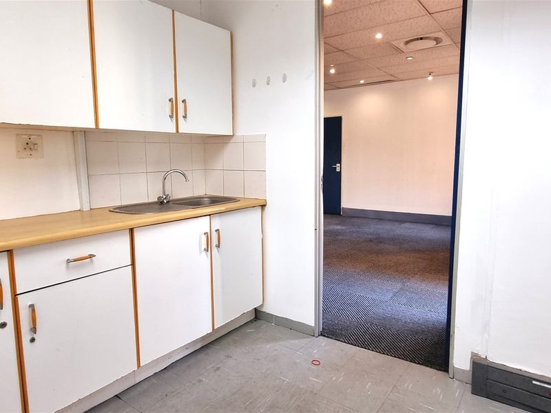 A functional office kitchenette featuring white cabinetry, a laminate countertop, and a stainless steel sink, leading into an open-plan office space with carpeted flooring.