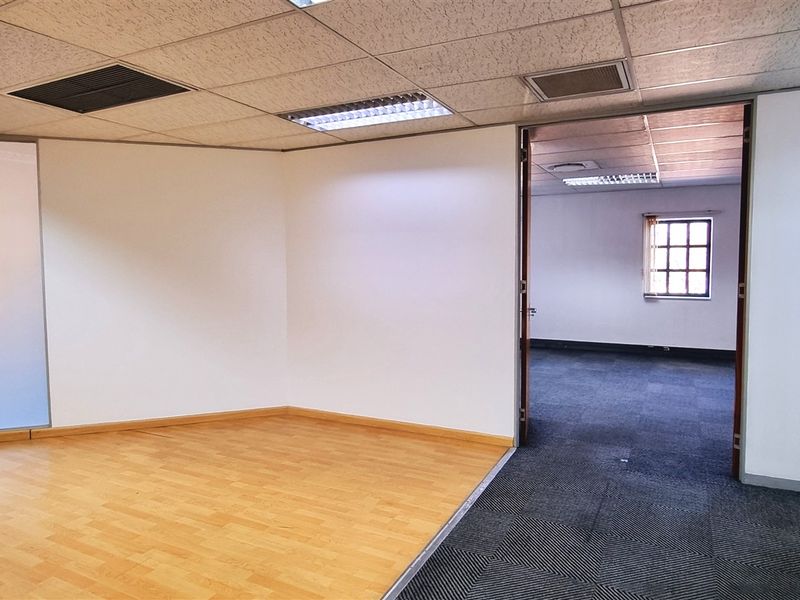 A bright, vacant office space featuring a mix of laminate and carpet flooring, suspended ceilings with recessed lighting, and a frosted glass partition.