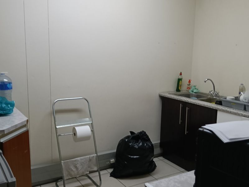 A functional office kitchenette featuring tiled flooring, laminate countertops, base cabinetry, and a wall-mounted air conditioning unit.
