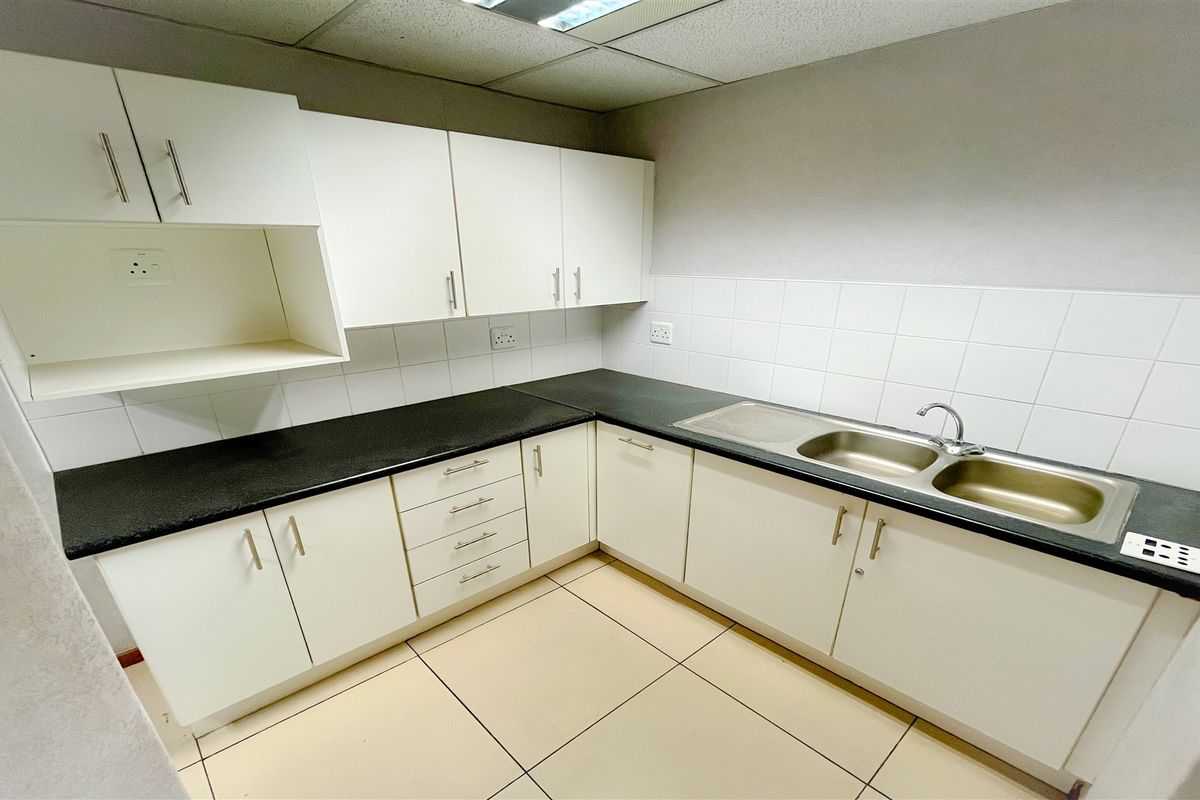 A clean and functional office kitchenette featuring white cabinetry, dark laminate countertops, a double stainless steel sink, and tiled flooring.
