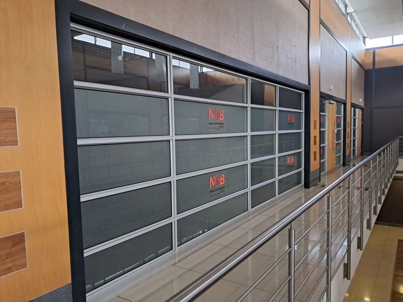 A modern office hallway featuring tiled flooring, wood veneer wall paneling, and a stainless steel balustrade overlooking the lower level.