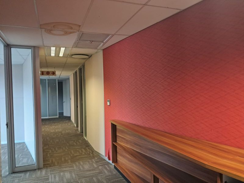 A professional office hallway featuring glass partitions, carpeted flooring, a built-in wooden shelving unit, and a distinctive red accent wall.