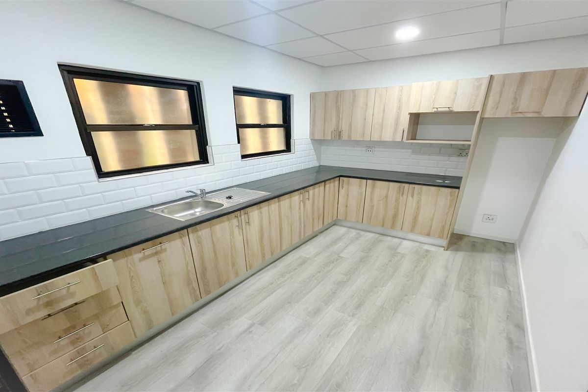 A modern office kitchenette featuring light wood-grain cabinetry, dark countertops, a stainless steel sink, and white subway tile backsplash.