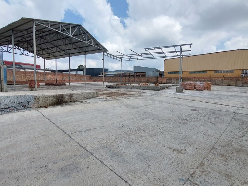 Expansive industrial yard featuring a large concrete surface, partial steel-framed roofing structures, and secure perimeter walling, suitable for storage or logistics operations.