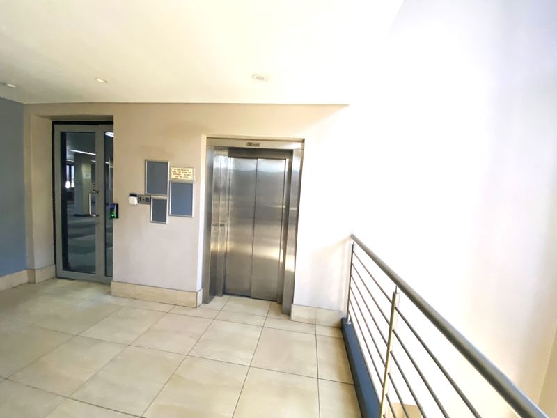 A clean and modern office building hallway featuring a stainless steel elevator, tiled flooring, and a secure glass entrance door.