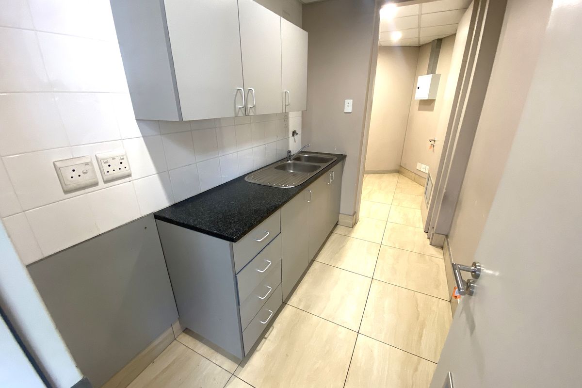 A clean, functional office kitchenette featuring built-in grey cabinetry, a stainless steel sink, dark countertops, and neutral tiled flooring.
