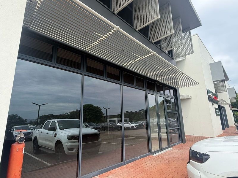 Modern retail storefront featuring extensive glass frontage, aluminium framing, and integrated sun shading elements, situated along a paved pedestrian walkway.