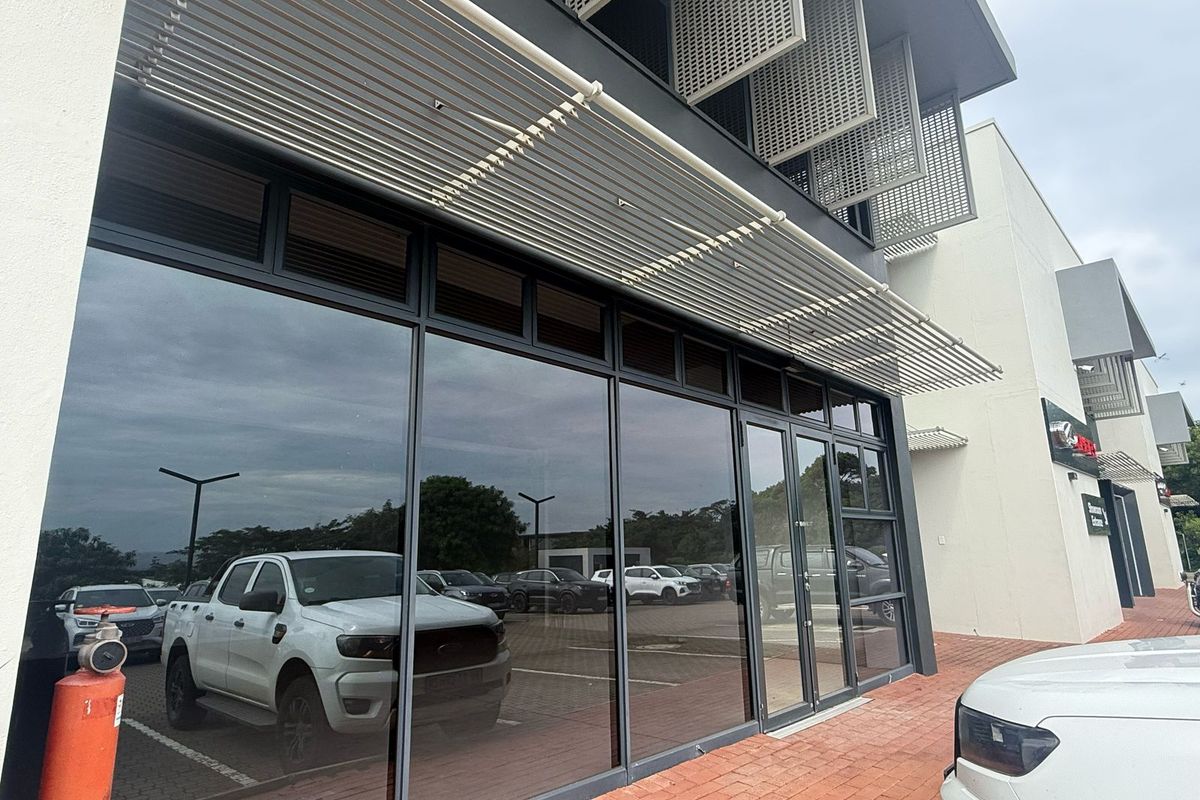 Modern retail storefront featuring extensive glass frontage, aluminium framing, and integrated sun shading elements, situated along a paved pedestrian walkway.