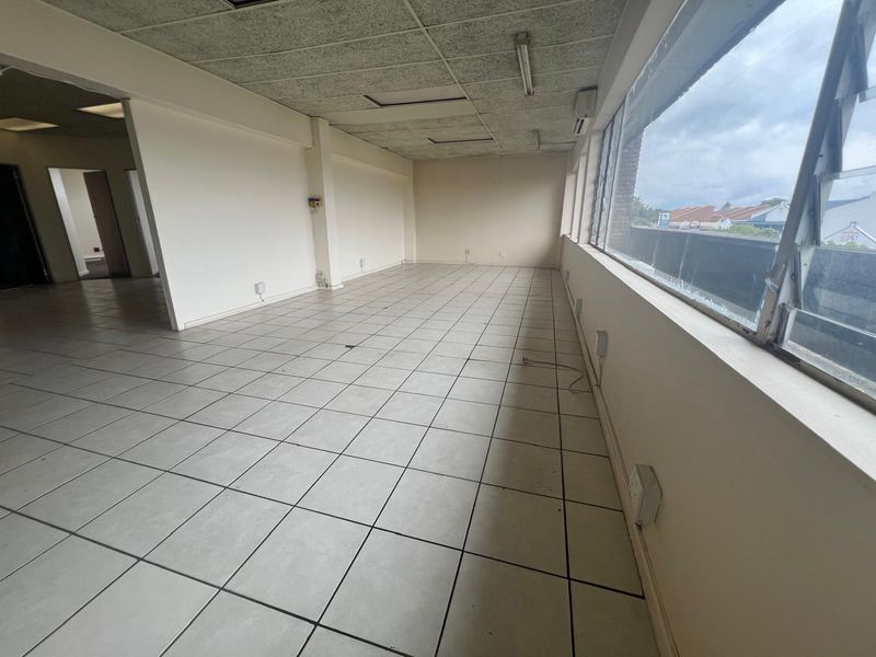An empty, open-plan commercial office space featuring tiled flooring, a suspended ceiling, and large windows providing natural light.