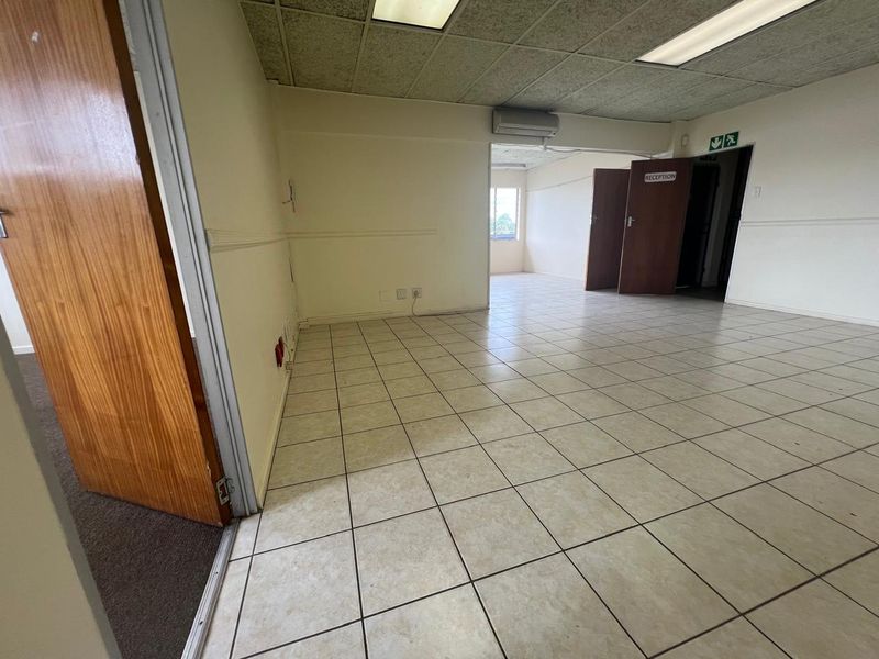 An empty commercial office space featuring ceramic tile flooring, a suspended ceiling with recessed lighting, and a wall-mounted air conditioning unit.