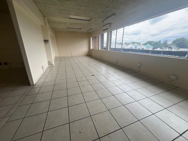 An empty commercial office space featuring tiled flooring, a suspended ceiling, and large windows providing natural light and exterior views.