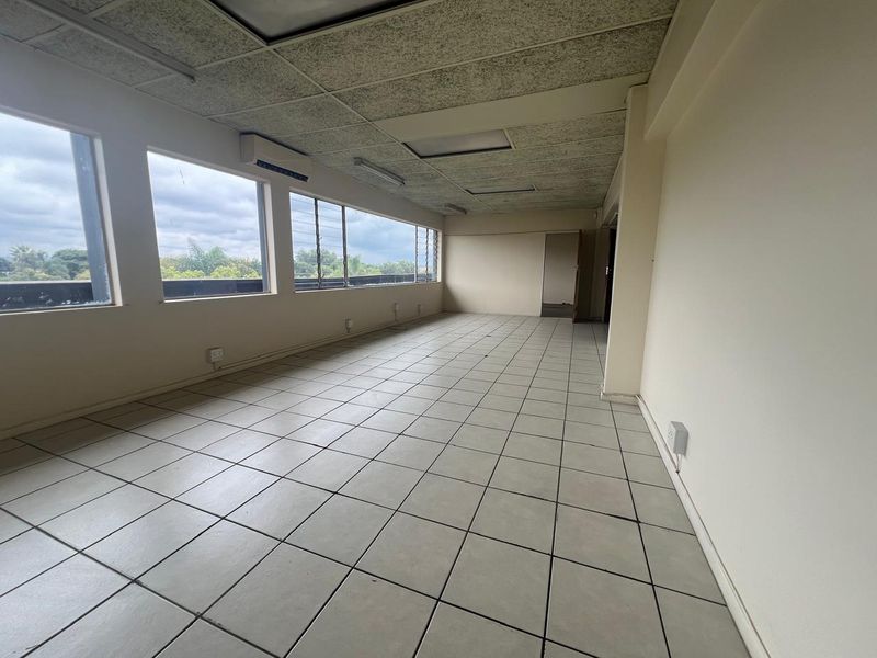 An empty, open-plan commercial office space featuring tiled flooring, a suspended ceiling, and large windows providing natural light.