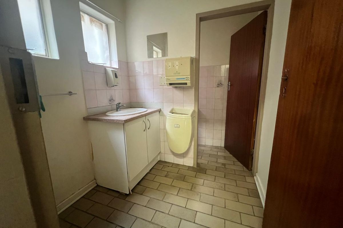 A functional commercial bathroom featuring a vanity cabinet with sink, tiled flooring, and wall tiling, presenting a dated aesthetic in need of modernization.