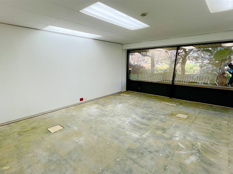 An empty office space featuring a suspended ceiling, recessed lighting, and large windows with garden views, currently requiring new flooring installation.