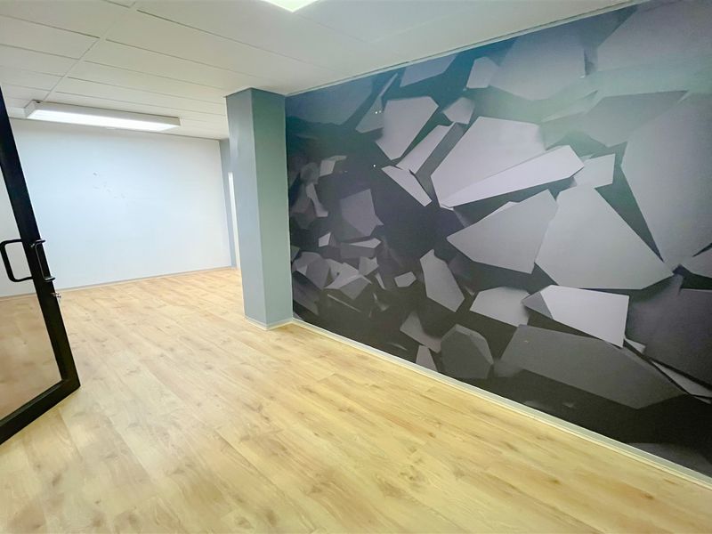 A bright, vacant office space featuring light wood-look laminate flooring, a suspended ceiling, and a modern geometric feature wall.