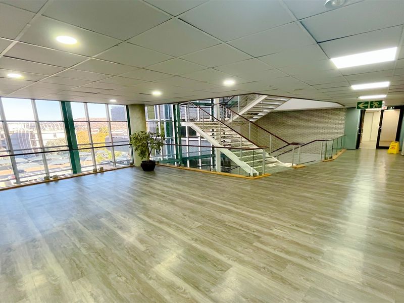 Spacious office building hallway featuring wood-look flooring, a large glass curtain wall with natural light, and an open staircase.