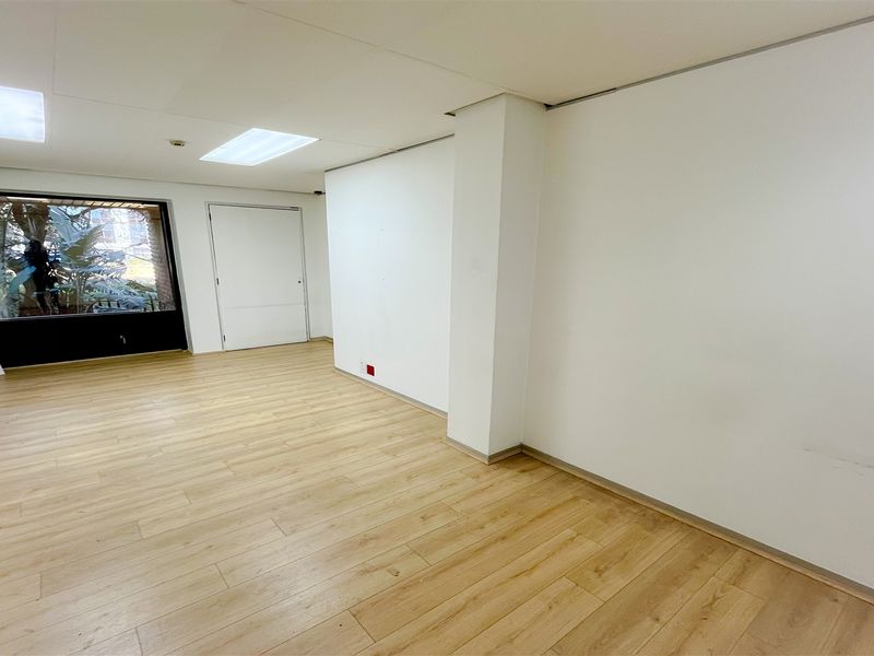 A bright, empty office space featuring modern laminate flooring, recessed ceiling lighting, and a large window providing natural light.