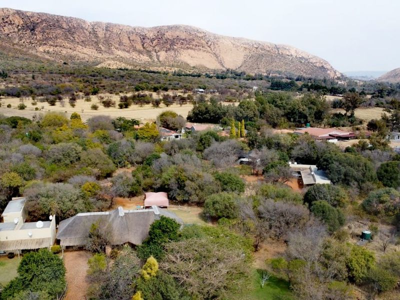An aerial view of a farm property featuring a thatched-roof dwelling set within a mature, tree-filled landscape against a scenic mountain backdrop.