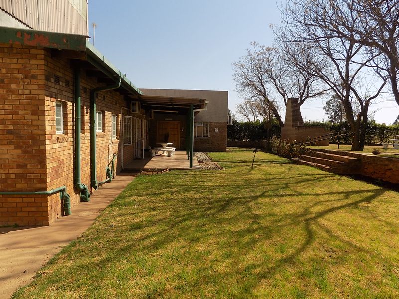 Rear exterior view of the commercial property featuring a brick facade, covered patio area, built-in braai, and landscaped garden space with stone steps.