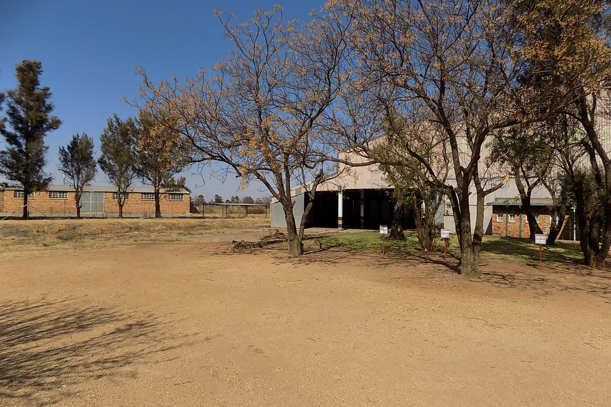 Rear exterior view of a commercial property featuring metal-clad structures, brick outbuildings, and a large unpaved yard area with mature trees.