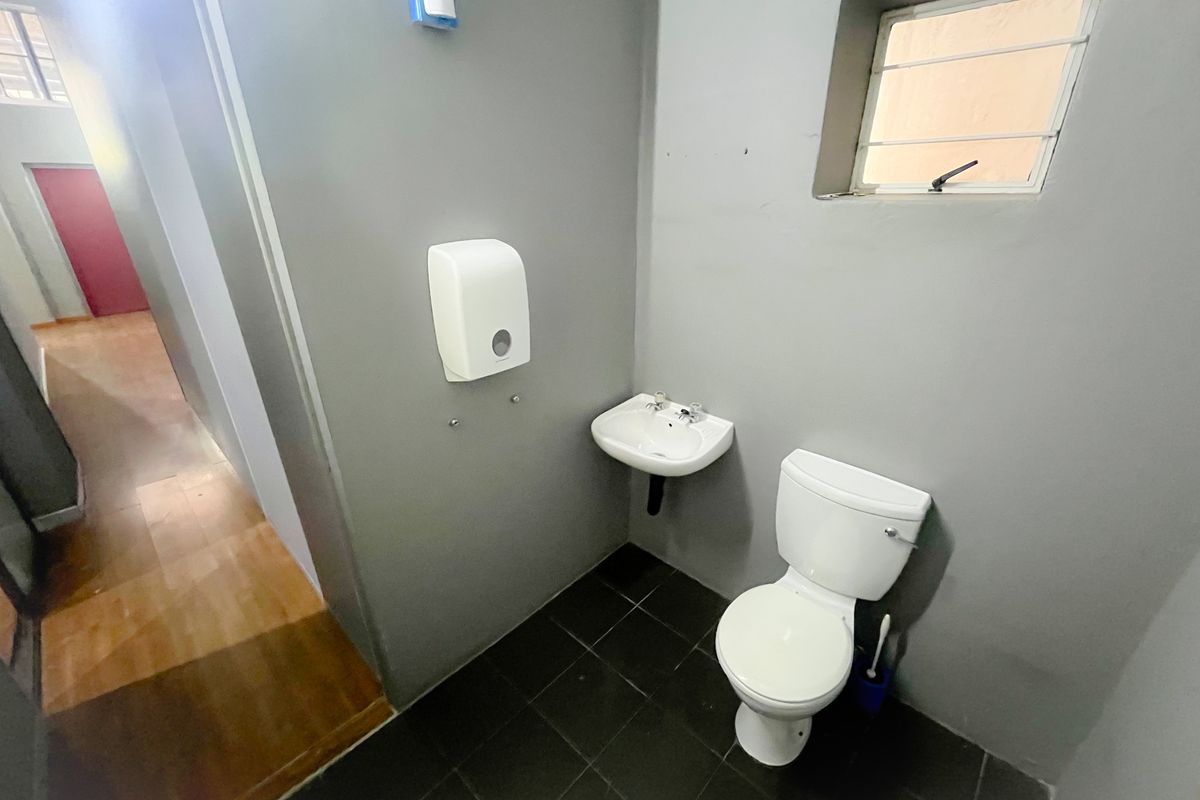 A functional office restroom featuring a wall-mounted basin, a standard toilet, and dark floor tiling with grey painted walls.