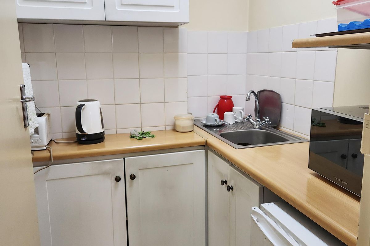 A compact kitchenette featuring white cabinetry, laminate countertops, a stainless steel sink, and tiled splashbacks.