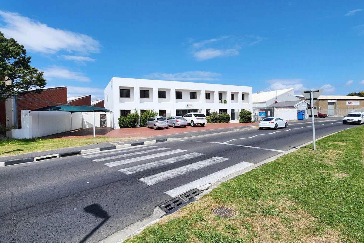 Street view of a commercial property featuring a white multi-story building with a paved parking area and perimeter wall.
