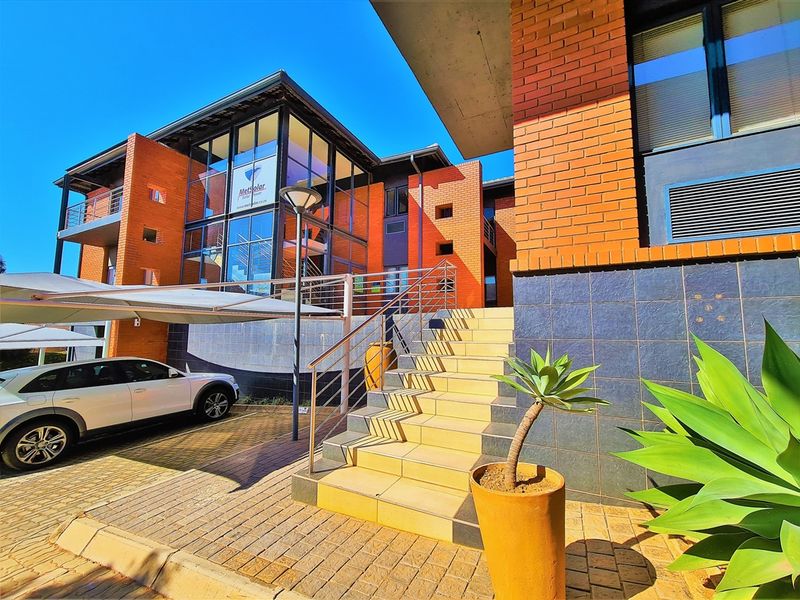 Modern office building exterior featuring a brick and glass facade, concrete staircase with stainless steel handrails, and a paved parking area with carport.
