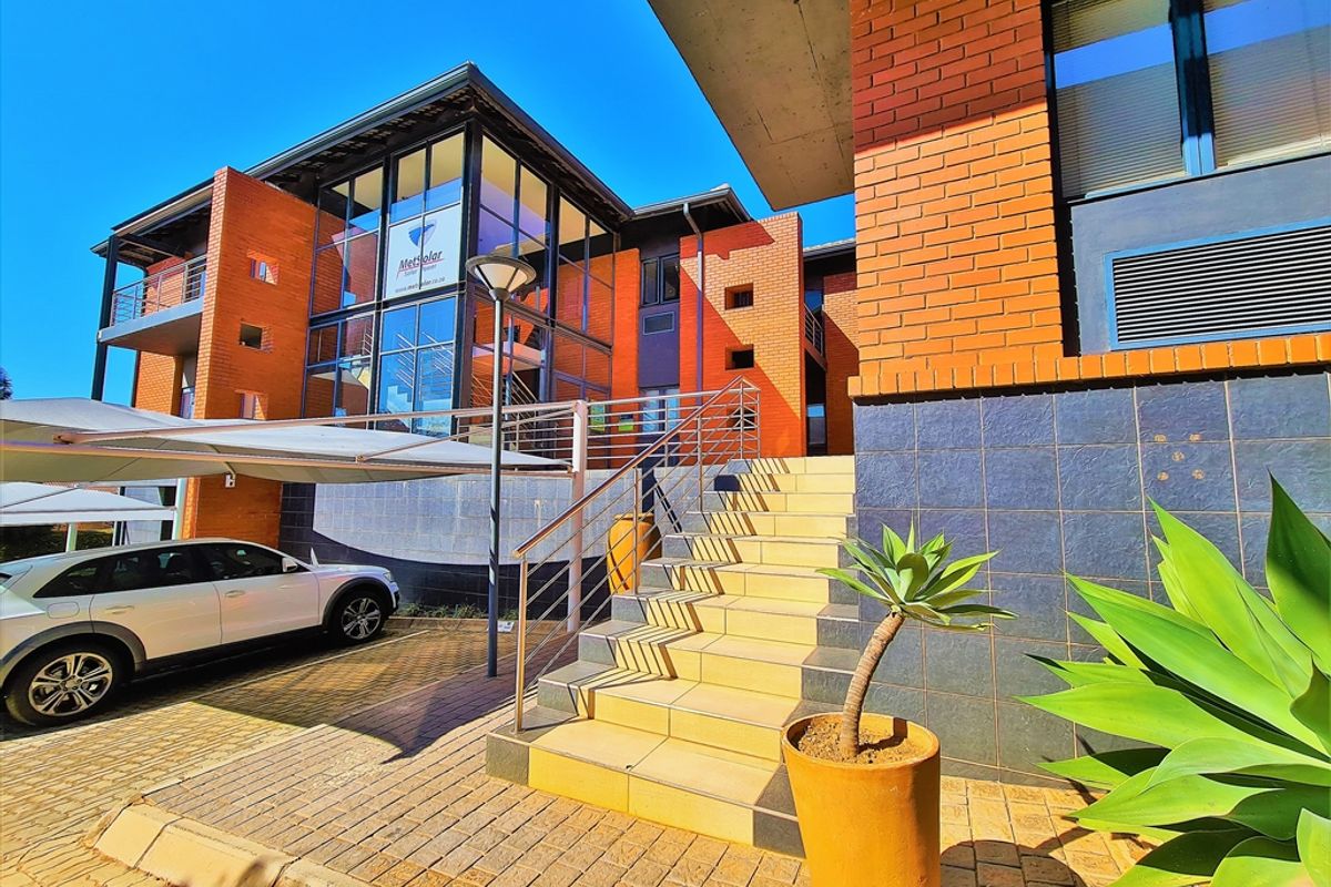 Modern office building exterior featuring a brick and glass facade, concrete staircase with stainless steel handrails, and a paved parking area with carport.