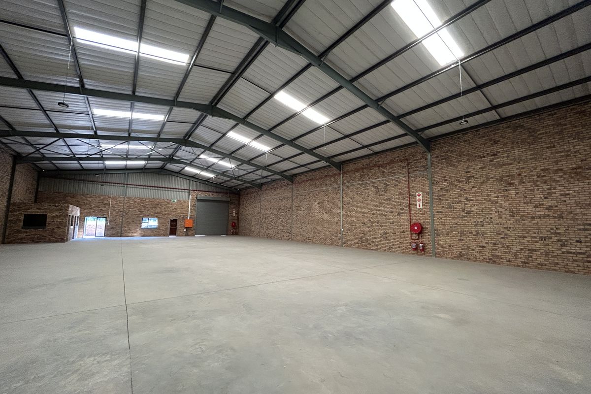 Spacious industrial warehouse featuring exposed brick walls, concrete flooring, high steel truss ceilings with integrated skylights, and essential fire safety equipment.