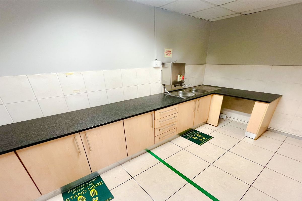 A functional office kitchenette featuring light wood cabinetry, dark laminate countertops, a stainless steel sink, and a wall-mounted hot water dispenser.