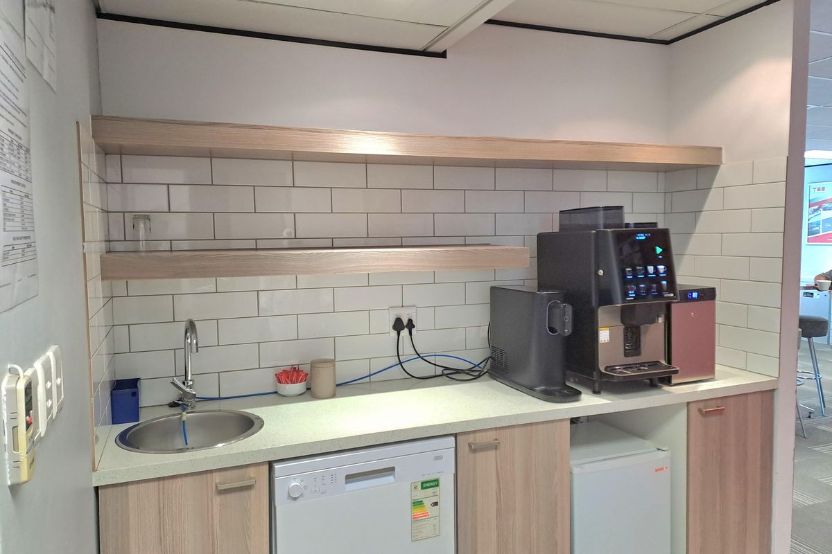 A compact office kitchenette featuring light wood cabinetry, white subway tile backsplash, floating shelves, a stainless steel sink, and integrated appliance space.