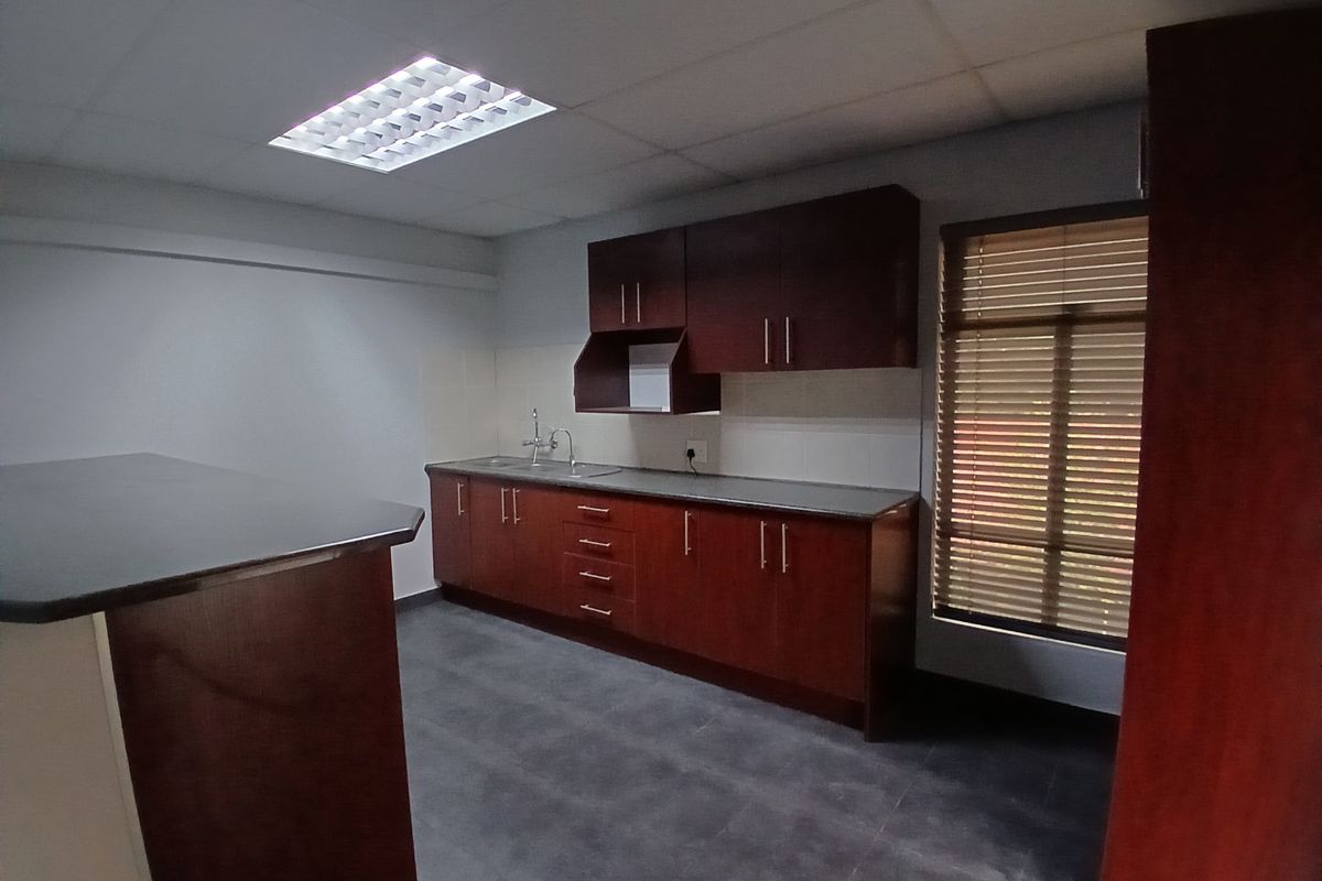 A functional office kitchenette featuring dark wood cabinetry, a countertop, a stainless steel sink, and a suspended ceiling with integrated lighting.