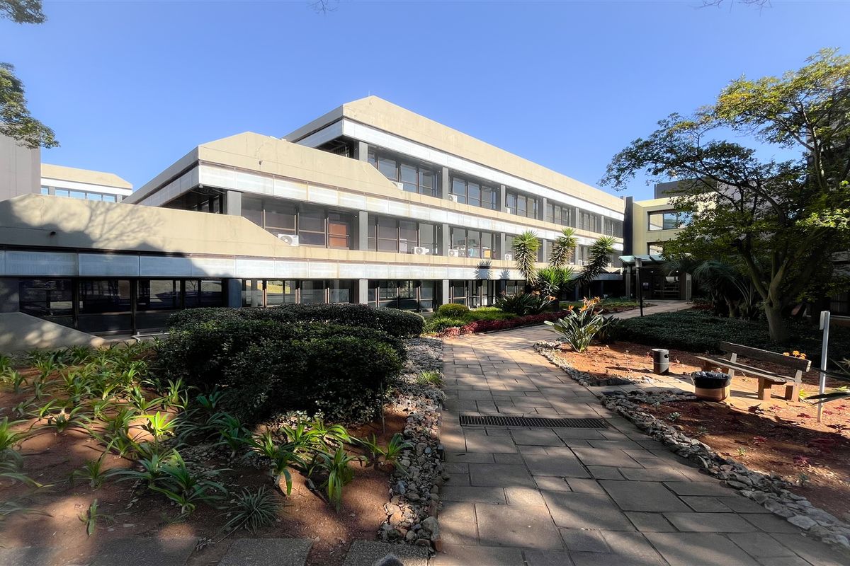 Exterior view of a multi-story office building featuring a concrete facade, large glass windows, and a paved walkway through landscaped garden grounds.