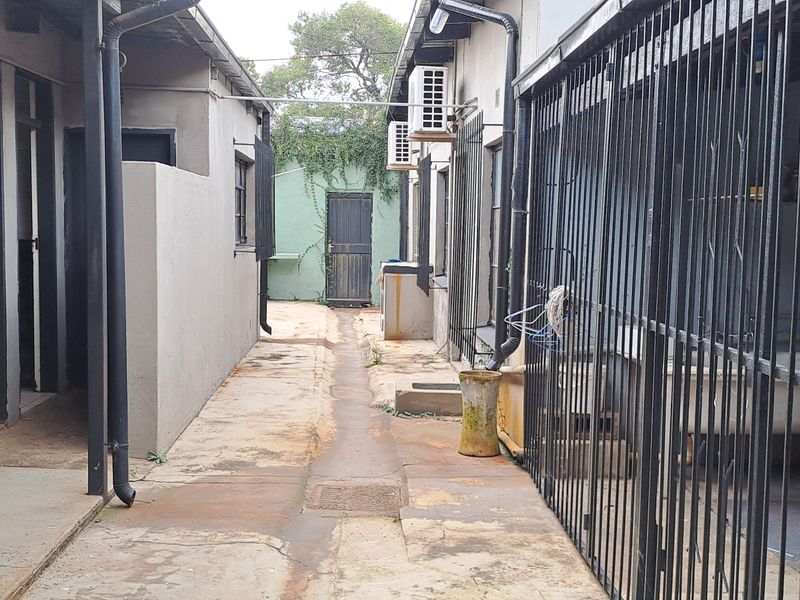 A narrow exterior rear walkway featuring concrete paving, security gates, and wall-mounted air conditioning units between two building sections.