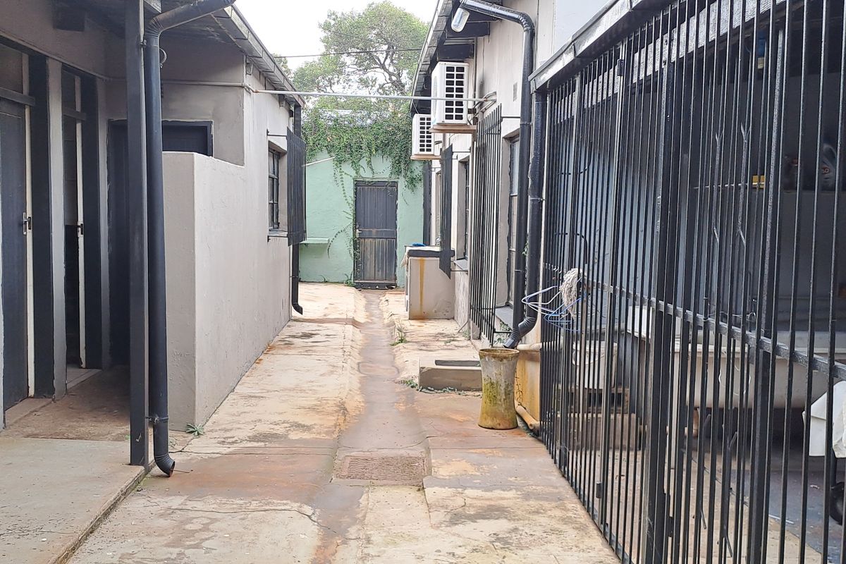 A narrow exterior rear walkway featuring concrete paving, security gates, and wall-mounted air conditioning units between two building sections.