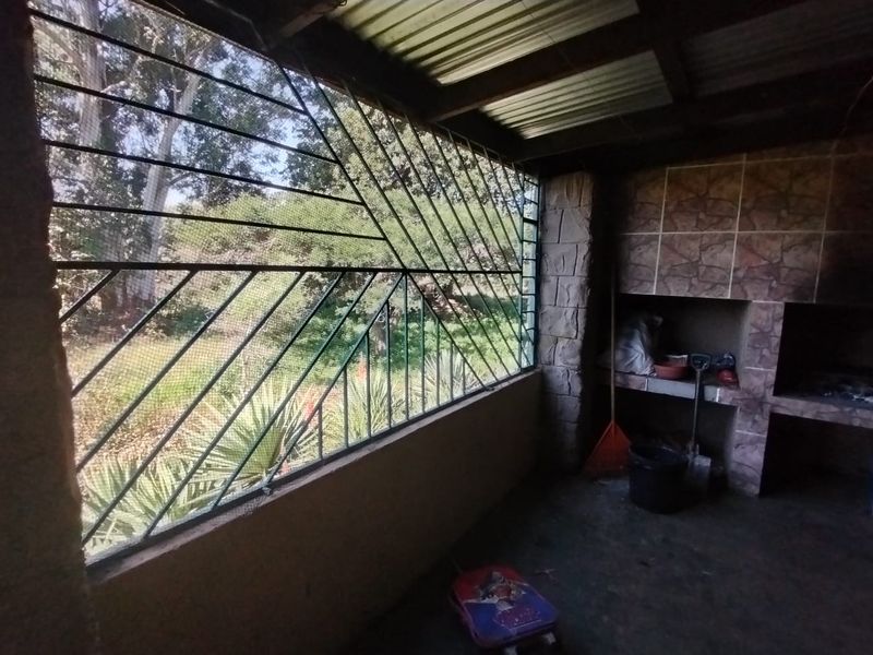 Covered outdoor patio area featuring a built-in stone-clad braai and security screening, overlooking the farm garden.