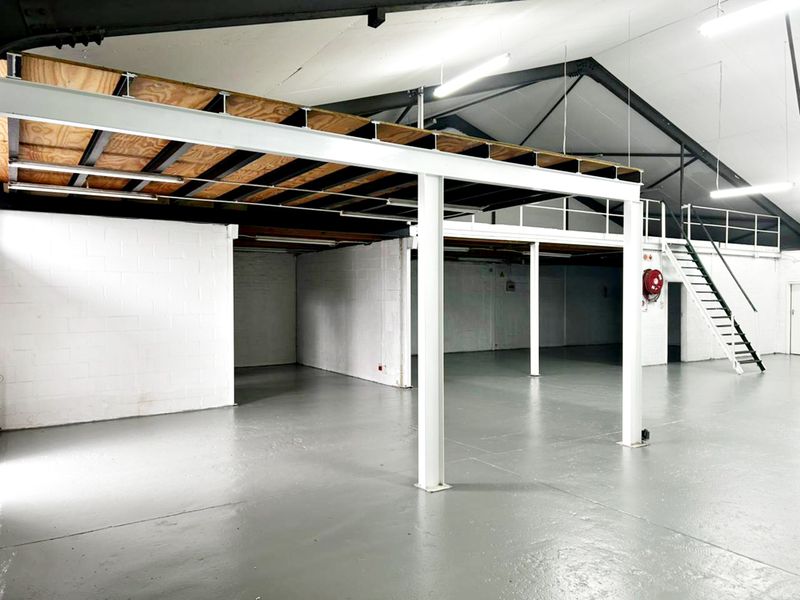 Spacious industrial warehouse unit featuring a mezzanine level, exposed steel roof trusses, painted concrete floors, and a dedicated steel staircase.