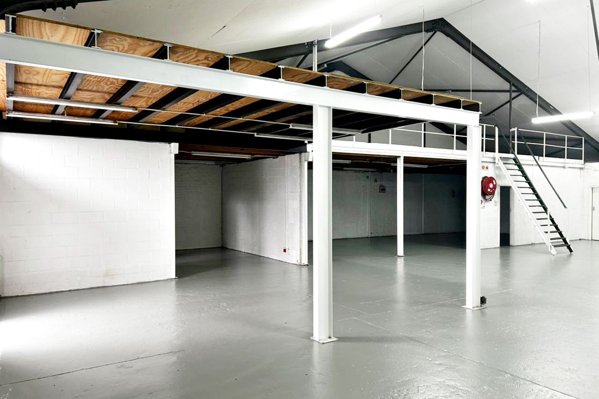 Spacious industrial warehouse unit featuring a mezzanine level, exposed steel roof trusses, painted concrete floors, and a dedicated steel staircase.