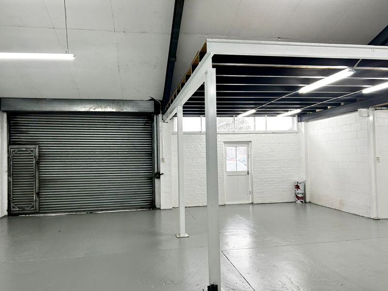 Spacious industrial utility area featuring a high roller shutter door, mezzanine level, painted concrete flooring, and bright overhead fluorescent lighting.