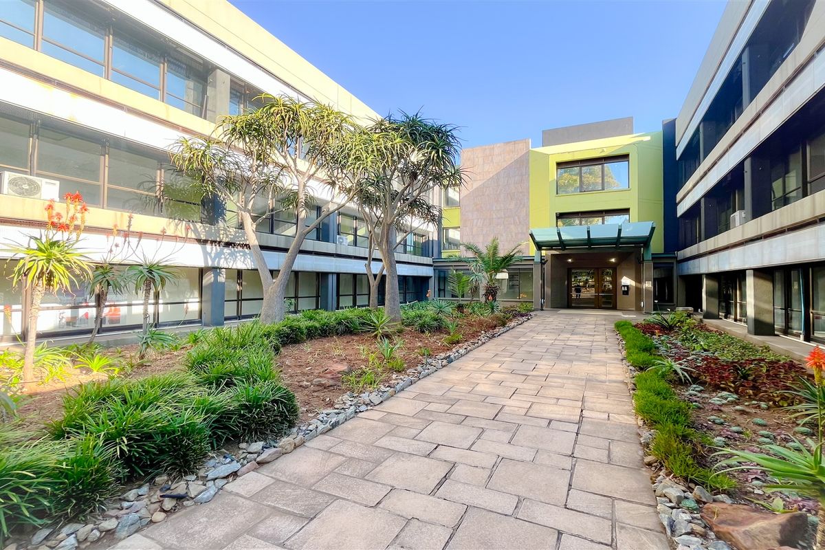 A well-maintained office building courtyard featuring a paved walkway, landscaped garden beds, and modern architectural facade with large windows.